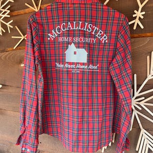 Può includere: Camicia a maniche lunghe a quadri rossi con la scritta "McCallister Home Security" e una grafica di una casa. La camicia ha anche la scritta "You're Never Home Alone" e "EST. 1990". La camicia è appesa a una gruccia di legno.