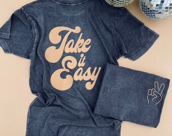 May include: A dark blue t-shirt with the words "Take it Easy" in a retro font on the back. The folded shirt shows a peace sign on the sleeve. Two disco balls are in the background.