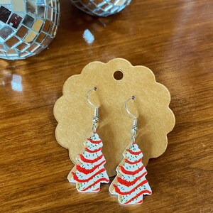 Puede incluir: Un par de pendientes de árbol de Navidad. Los pendientes son blancos con rayas rojas y chispas verdes. Los pendientes cuelgan de ganchos plateados. Los pendientes están sobre una tarjeta marrón.