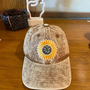 May include: A brown baseball cap featuring a yellow sunflower design. The sunflower has a black, patterned centre. The cap has a curved peak and a button on top. The cap is made of a faded, textured material. The cap is a stylish accessory.
