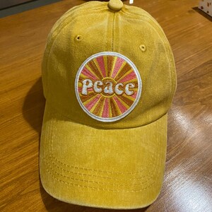 May include: A mustard yellow baseball cap with a circular patch that says "peace" in a retro font. The patch has a pink and orange sunburst design.