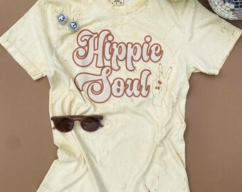 May include: Cream-colored t-shirt with the words "Hippie Soul" in brown script and a peace sign hand graphic. The shirt has a vintage, distressed look. Sunglasses and disco ball earrings are also in the image.