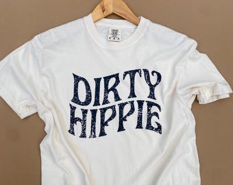 May include: A white short-sleeve t-shirt featuring the text DIRTY HIPPIE in a dark navy blue, distressed, wavy font across the chest. The shirt has a crew neckline and a Comfort Colors brand label visible at the inner collar, hanging on a wooden hanger.