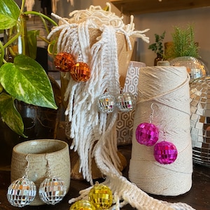 Puede incluir: Surtido de pendientes de bola de discoteca en varios colores, incluyendo plata, oro, naranja y rosa. Los pendientes se muestran sobre una superficie de madera con hilo blanco y una taza de cerámica.