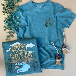 Può includere: Una maglietta blu turchese con una scena di bosco e il testo "It's all good in the woods" stampato sul davanti. La maglietta è piegata e appoggiata su una superficie marrone con due ornamenti di ceramica a forma di fungo.