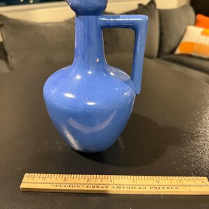 May include: A blue ceramic pitcher with a handle and a small spout. The pitcher has a rounded body and a textured surface. A wooden ruler is in the foreground.
