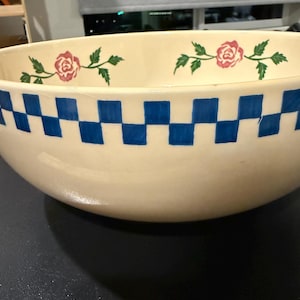 May include: A large cream-colored ceramic bowl featuring a blue checkered pattern around the upper exterior rim and a painted pink rose and green leaf motif along the interior edge, positioned next to a ruler showing a measurement of 4 inches.