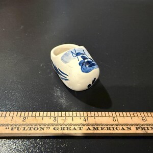 May include: A small, white ceramic clog with blue floral and line designs. The miniature shoe is a traditional Dutch souvenir. The clog is approximately 2 inches long.
