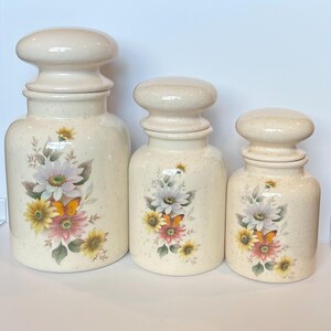 May include: Set of three ceramic canisters with floral and butterfly designs. The canisters are white with a speckled finish and feature a light pink, yellow, and orange floral pattern with a butterfly on each.
