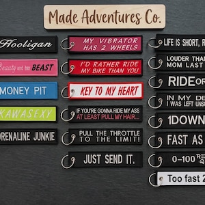May include: A collection of rectangular fabric keychains with metal rings, featuring various embroidered phrases like Hooligan, Money Pit, and Ride or Die. The brand name Made Adventures Co. is printed on a wooden sign above the keychains.