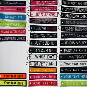 Könnte beinhalten: Eine Sammlung rechteckiger Schlüsselanhänger aus Stoff mit gesticktem Text in verschiedenen Farben wie Schwarz, Pink, Blau, Grün, Rot, Orange und Weiß. Jeder Anhänger verfügt über eine Metallöse und einen Schlüsselring zur Befestigung.