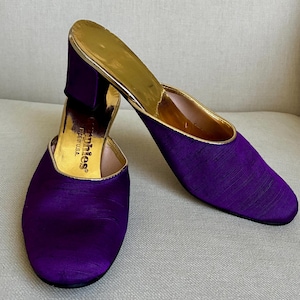 May include: A pair of purple satin mules with a gold-colored interior and heel. The shoes have a rounded toe and a block heel. The brand name "Sophies" is visible inside the shoe. Made in the USA.