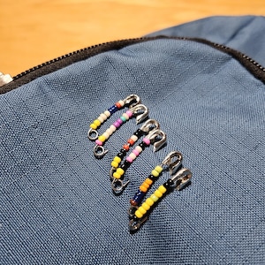 May include: Close-up of four beaded safety pins. Each pin features a different color pattern of small beads, including pink, yellow, black, and blue. The pins are silver-toned and are arranged on a blue fabric surface.