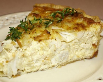 May include: A portion of quiche on a white plate with a decorative edge. The quiche features a golden-brown pastry base and a creamy, white filling. Fresh green herbs are used as a garnish on top of the quiche.