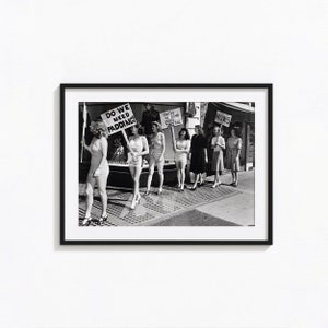 Może przedstawiać: Czarno-białe zdjęcie grupy kobiet trzymających tabliczki z napisami "Do We Need Padding?", "United We Stand, Divided We Fall" i "Now's". Kobiety są ubrane w stroje kąpielowe i stoją na miejskiej ulicy.