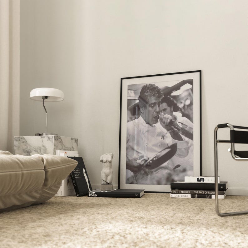 May include: A black and white photograph of a chef cooking in a kitchen. The photo is framed in a black frame and is hanging on a white wall. There are books and a sculpture on a table below the photo.