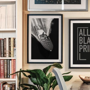 May include: Black and white framed print of a speedboat racing across dark water, creating a white wake. The boat has a classic design with two figures on board. The print is displayed on a wall with other framed art and a bookshelf.