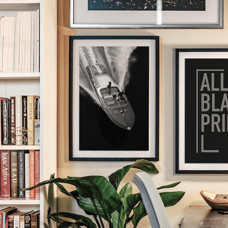 May include: Black and white framed print of a speedboat racing across dark water, creating a white wake. The boat has a classic design with two figures on board. The print is displayed on a wall with other framed art and a bookshelf.