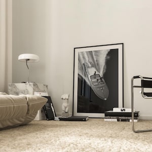 May include: A black and white framed print of a speedboat racing across the water. The print is displayed in a room with a marble side table, a white lamp, books, and a small sculpture. The room has a neutral color palette.