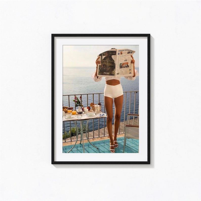 May include: A framed print featuring a person holding a newspaper, standing on a balcony overlooking the ocean. A small table with breakfast items is set up nearby. The person is wearing white shorts and sandals. The print has a black frame.