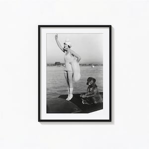 May include: Black and white framed photograph of a woman in a vintage swimsuit and cap, standing on a boat and pointing upwards. A dog sits nearby, also on the boat. The background shows water and distant buildings.