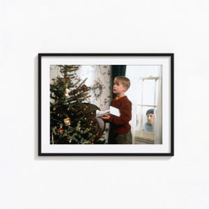 May include: A framed print featuring a young boy in a red sweater holding a book near a decorated Christmas tree. An older man is visible through a window in the background. The scene evokes a holiday theme.