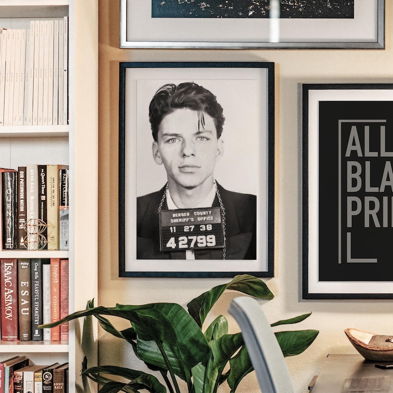 May include: A black and white photo of a man in a mugshot. The photo is framed in a black frame and hanging on a wall. The man is wearing a chain around his neck and has a number tag on his chest that reads "42799".