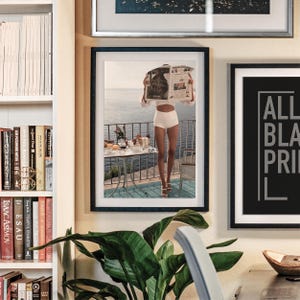 May include: Framed print of a person on a balcony reading a newspaper, with a table set for breakfast. The person is wearing white shorts and a white top. The background shows a seascape. Other framed art and a bookshelf are also visible.