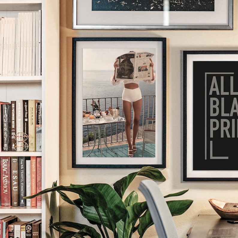 May include: Framed print of a person on a balcony reading a newspaper, with a table set for breakfast. The person is wearing white shorts and a white top. The background shows a seascape. Other framed art and a bookshelf are also visible.