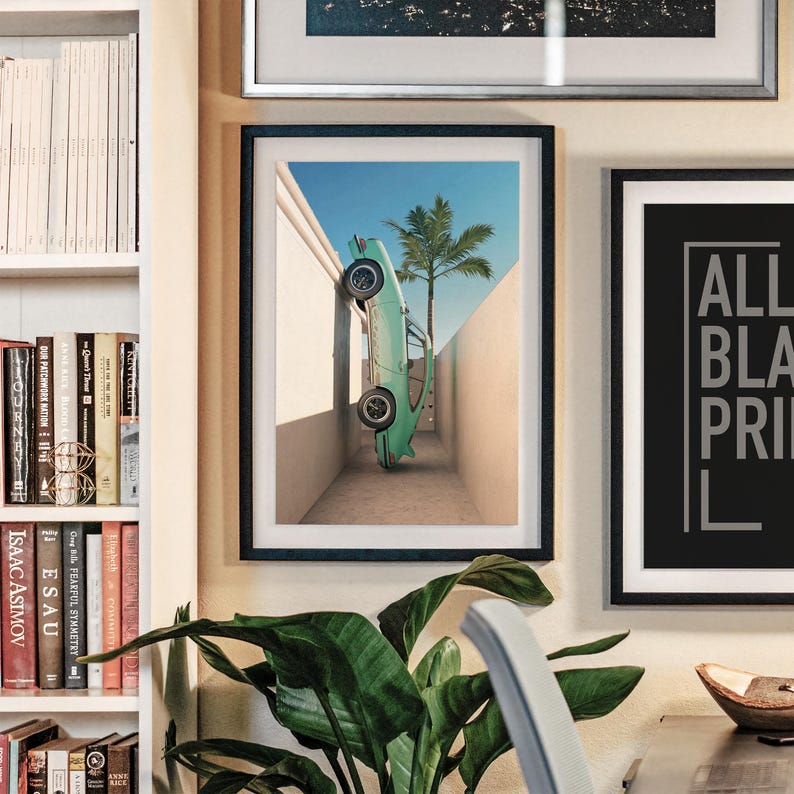 May include: A framed art print featuring a vintage teal car leaning against a wall with a palm tree in the background. The artwork is displayed on a wall with other framed pieces and a bookshelf.