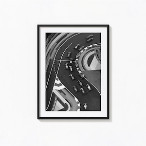 May include: Black and white framed print of a Formula 1 race. The aerial view shows race cars speeding around a curved track. The track has white and gray markings. The print is in a black frame and is displayed on a white wall.