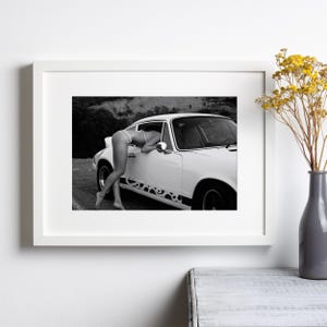 Op de afbeelding: Een zwart-witfoto van een vrouw in bikini die in een witte Porsche 911 Carrera leunt.