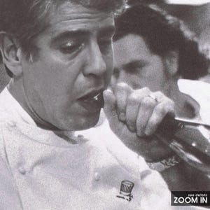 May include: A black and white photo of a man in a white chef's coat speaking into a microphone. He is wearing a chef's hat pin on his coat. The man is looking at the camera and smiling.