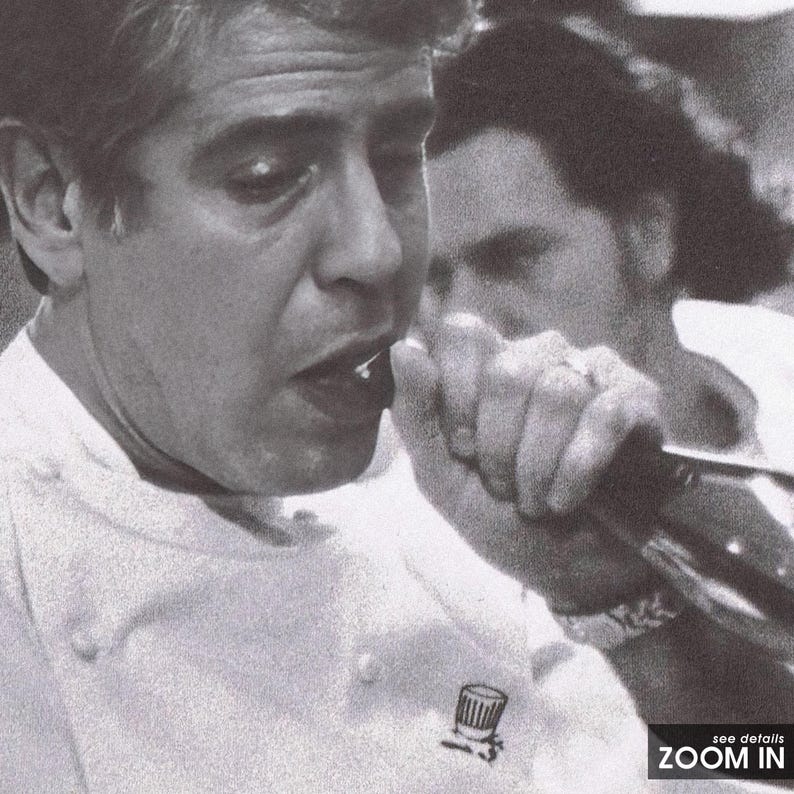 May include: A black and white photo of a man in a white chef's coat speaking into a microphone. He is wearing a chef's hat pin on his coat. The man is looking at the camera and smiling.