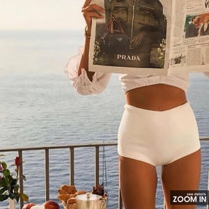 May include: A person wearing a white top and shorts, holding a magazine with the Prada logo. The scene is set on a balcony overlooking the ocean. A table with a basket of pastries and a silver tea set is visible.