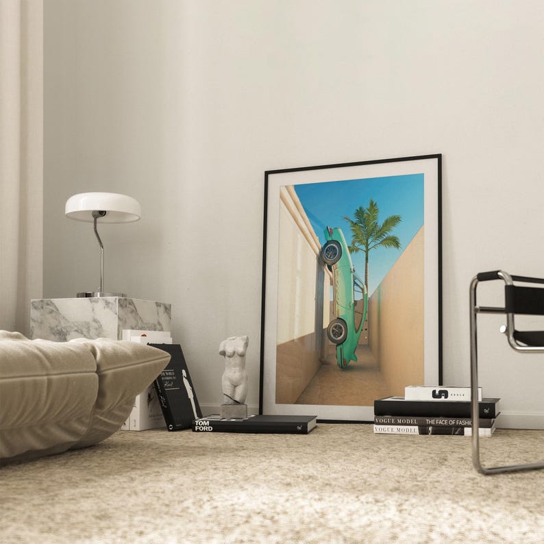 May include: A framed art print featuring a turquoise car leaning against a wall with a palm tree under a blue sky. The print is displayed in a room with a marble side table, a lamp, books, and a sculpture.