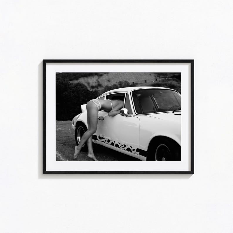 Op de afbeelding: Een zwart-witfoto van een vrouw in een witte bikini die in een witte Porsche 911 Carrera leunt.