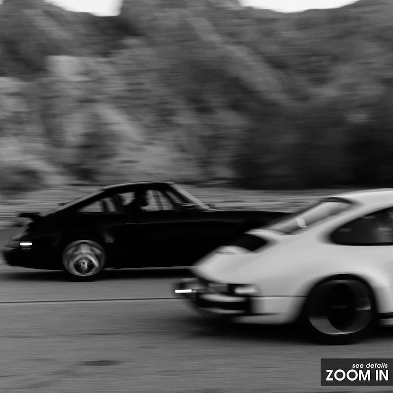 Puede incluir: Imagen en blanco y negro de dos coches deportivos cl&aacute;sicos en movimiento. El coche negro est&aacute; en primer plano, con el coche blanco ligeramente detr&aacute;s. El fondo est&aacute; borroso, sugiriendo velocidad.