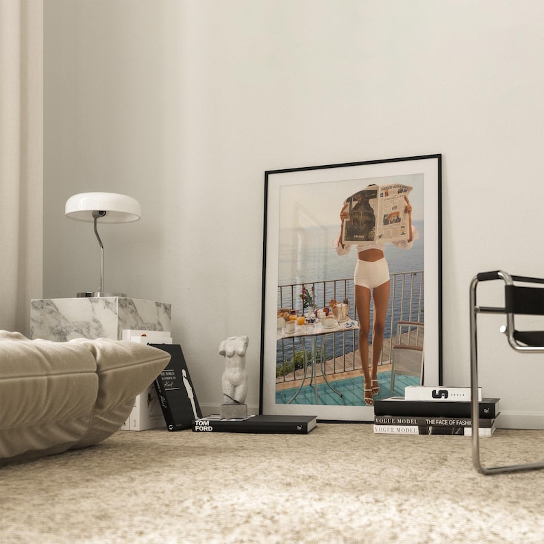 May include: A framed art print featuring a person holding a newspaper, standing on a balcony overlooking the sea. A white lamp, marble block, books, and a small sculpture are in the foreground. The room has a neutral color palette.