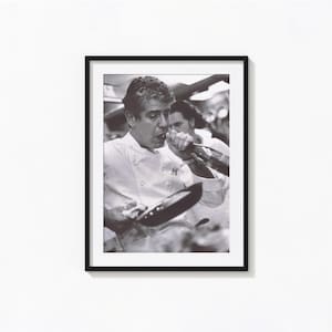 May include: A black and white photograph of a chef in a white chef's coat, holding a frying pan and drinking from a bottle.