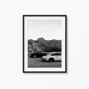 May include: Black and white framed print featuring two classic sports cars in motion on a road. The cars are a black coupe and a white coupe, with a blurred mountain range in the background. The image is a study in motion and contrast.