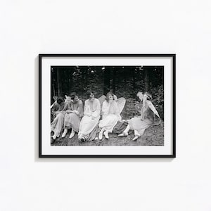 Op de afbeelding: Een zwart-witfoto van vijf vrouwen gekleed in witte gewaden met vleugels, zittend in een bosrijke omgeving. Ze kijken allemaal naar de camera en glimlachen.