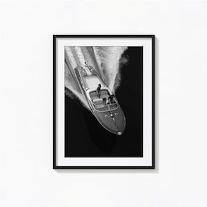 May include: Black and white framed print of a speedboat gliding across dark water, creating a wake. The boat has a wooden hull and is viewed from above. Two figures are visible on the boat. The print is displayed on a white wall.