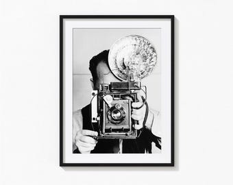 Op de afbeelding: Zwart-wit ingelijste print van een fotograaf met een vintage camera met flitser. De fotograaf draagt een vest en een wit overhemd. De lens en bedieningselementen van de camera zijn duidelijk zichtbaar.