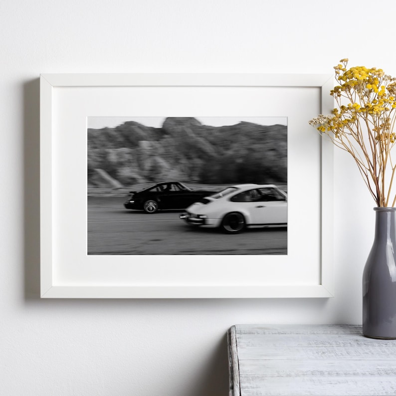 Puede incluir: Impresi&oacute;n en blanco y negro enmarcada que muestra dos coches deportivos en movimiento. Uno es negro, el otro blanco. El fondo muestra una cordillera borrosa, sugiriendo velocidad. La impresi&oacute;n est&aacute; enmarcada en blanco.