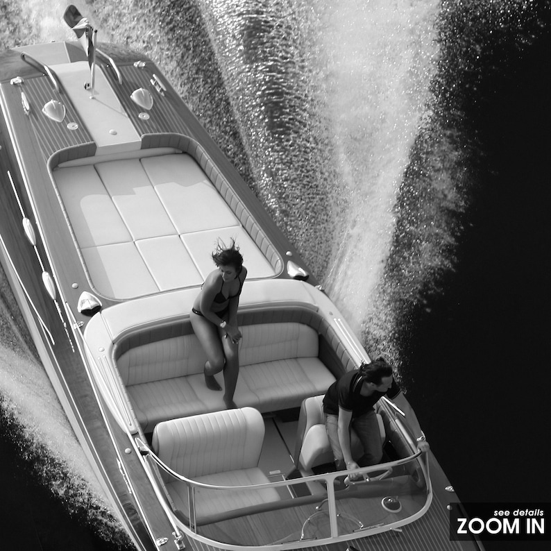 May include: Black and white image of a wooden speedboat in motion, with two people on board. The boat has white cushioned seating and is creating a large wake in the water. The text "ZOOM IN" is visible in the bottom right corner.