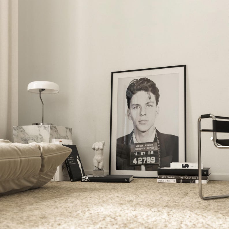 May include: A black and white photograph of a man in a mugshot, framed in a black frame, hanging on a white wall. The photo is surrounded by books and a small white sculpture.