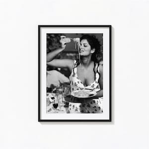 May include: A black and white photograph of a woman in a floral dress eating spaghetti with a fork. She is smiling and looking at the camera.