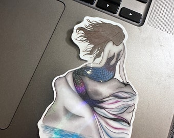 May include: A sticker of a mermaid with long flowing brown hair, a blue and green tail, and a white and pink tail fin. The mermaid is sitting on a rock with water below her.