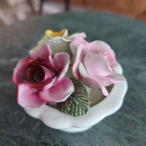 May include: A white porcelain figurine of a flower arrangement with pink, red, and yellow roses. The roses are arranged in a small white bowl.